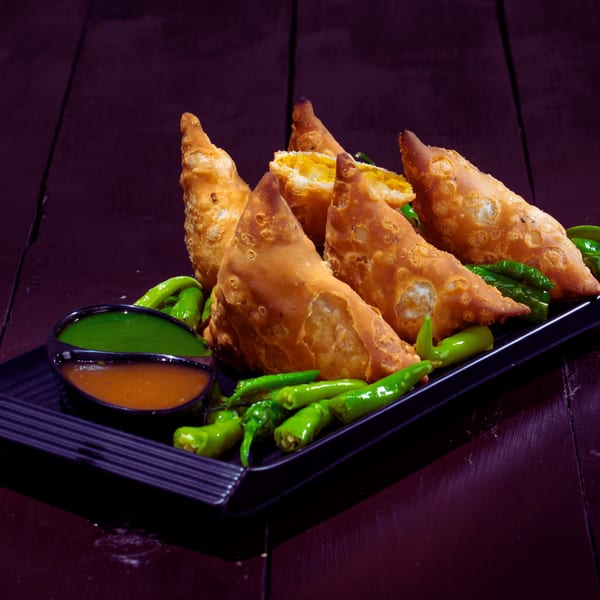 Golden-fried Indian samosas with green chili peppers and two dipping sauces on a black plate.